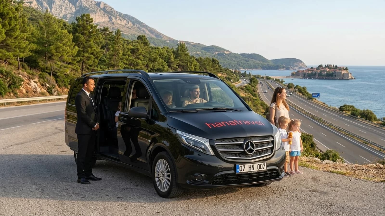 A black transfer van travelling along a scenic coastal road between the mountains and the Mediterranean Sea.