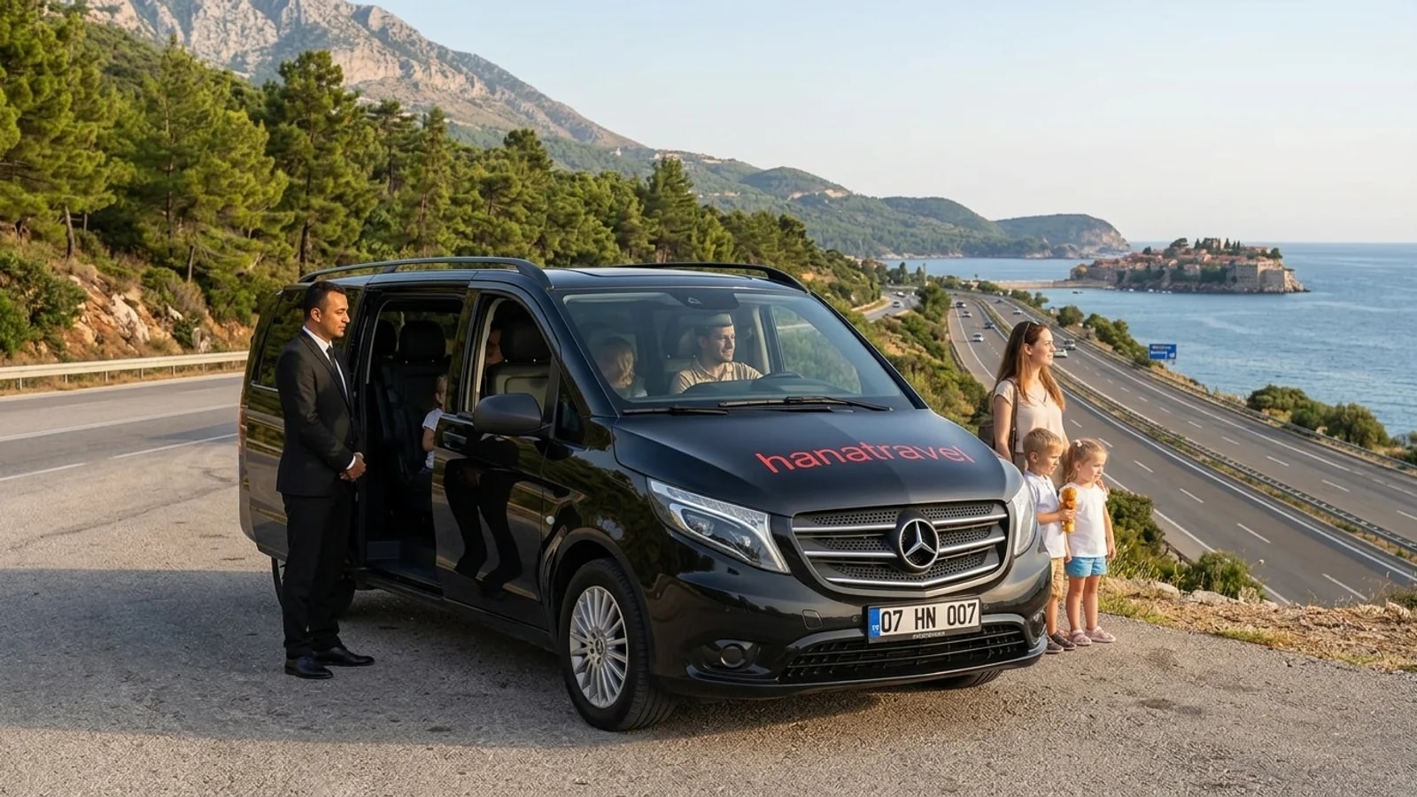 A black transfer van travelling along a scenic coastal road between the mountains and the Mediterranean Sea.