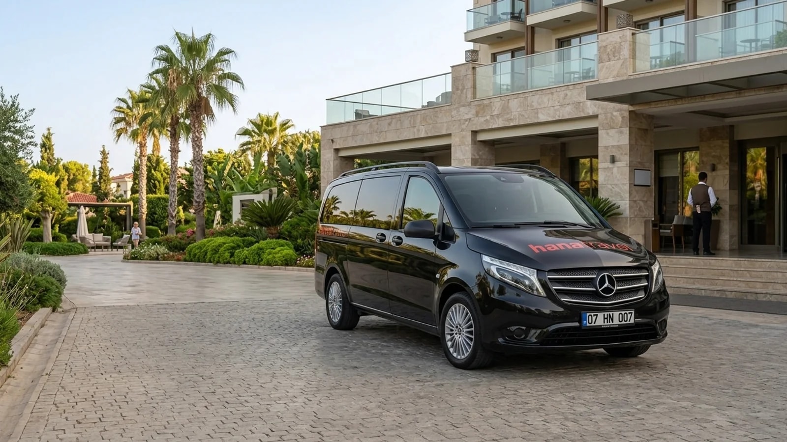 A black transfer van stands on a palm-lined driveway in front of a modern resort in Antalya.